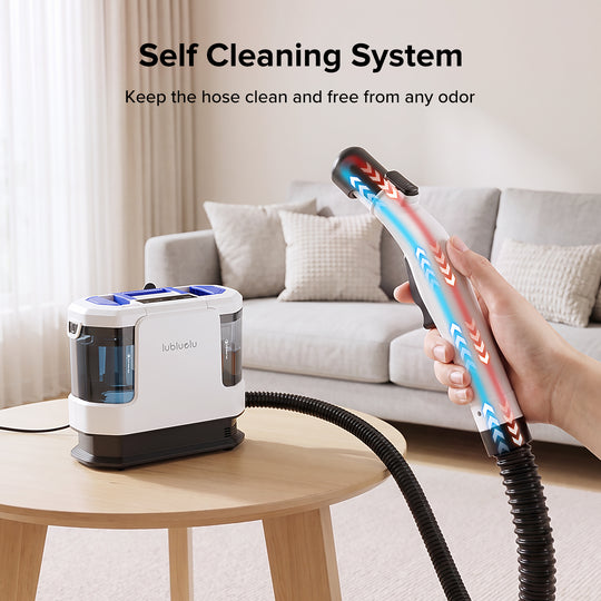 A handheld steam cleaner with a hose connected to a white and black base unit sits on a wooden table in a living room setting. A hand holds the steam nozzle, which displays red and blue steam patterns.