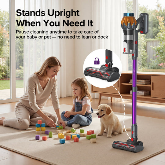 A cordless stick vacuum cleaner with a purple handle and orange and gray upper section stands upright on a light rug in a bright living room. A woman and a young girl are sitting on the floor playing with blocks, and a small dog is sitting nearby.